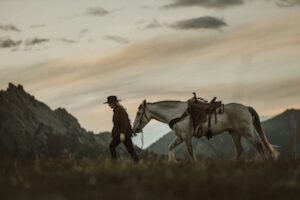 A woman in a fringed leather jacket with cowboy hat leading a grey horse with a Western saddle through the American countryside